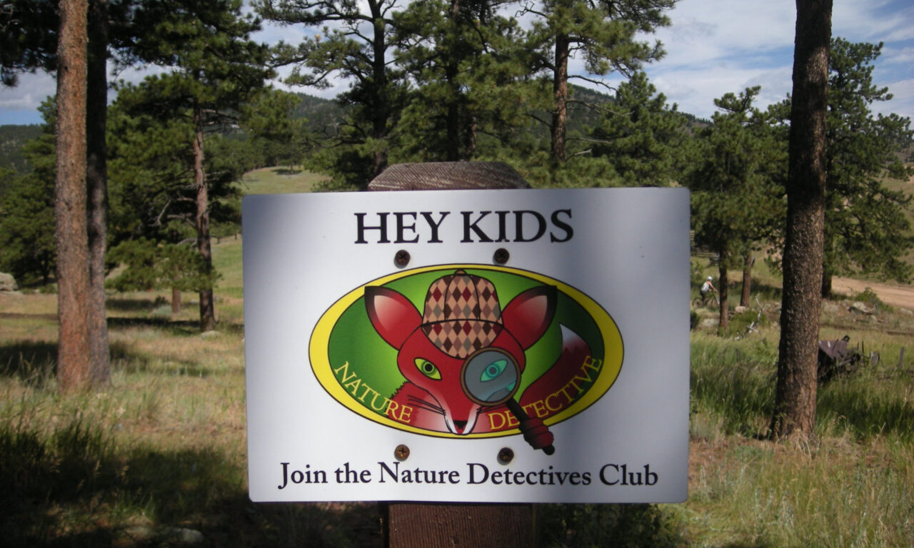 A park sign featuring an illustration of Sherlock Fox, wearing a detective hat and holding a magnifying glass, invites kids to 