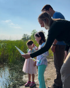 Two small girls stand with their parents at the edge of a pond. The Mother holds a mystery guide in her hand. They are working as a team to complete the guide.