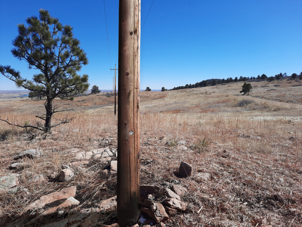 A landscape showing a re-creation of what the area looked like before the removal of a utility pole. The foreground features a tall wooden pole standing among dry grass and scattered rocks. In the background, there are rolling hills dotted with pine trees under a clear, bright blue sky. The scene is open and expansive, with more utility poles visible in the distance, marking the previous path of the power line.