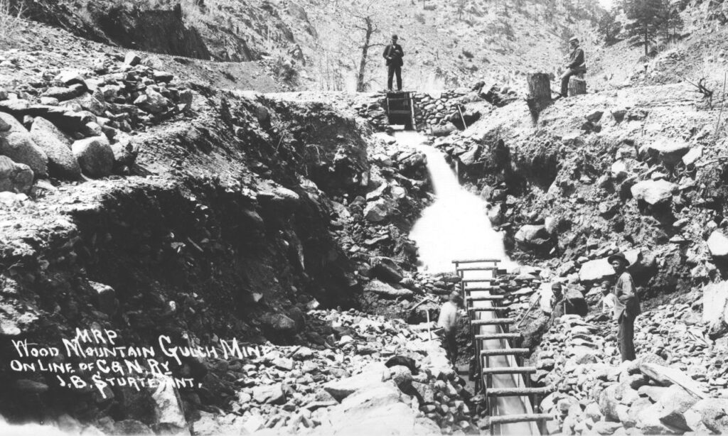 A group of miners work alongside a rocky streambed at a placer mine, with a wooden sluice running through the center of a rugged canyon.