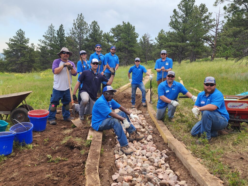 A group of volunteers in blue shirts and work gloves pose together while constructing a rock-lined trail in a scenic, forested area with tall pine trees and grassy meadows.