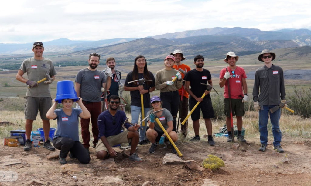 A group of Verily employees pose with trail-building tools on a rocky hillside with scenic mountains in the background.