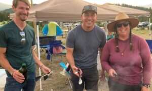 Three smiling people stand in front of a Camp Chef grill cooking burgers and chicken at an outdoor campsite.