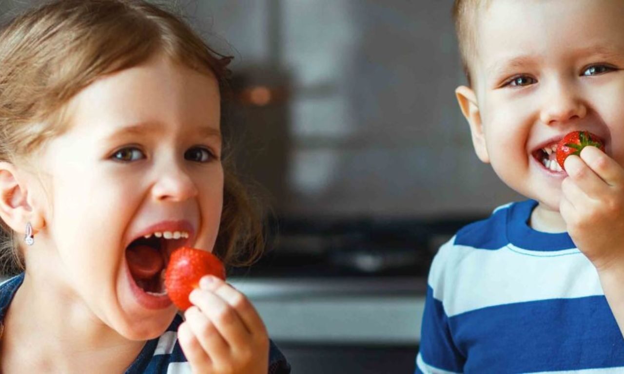 young girl and young boy both enjoy a ripe strawberry with big smiles on their faces.