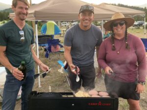 Three smiling people stand in front of a Camp Chef grill cooking burgers and chicken at an outdoor campsite.