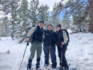 Three uniformed rangers smile and pose together while snowshoeing on a snowy trail surrounded by snow-covered pine trees.