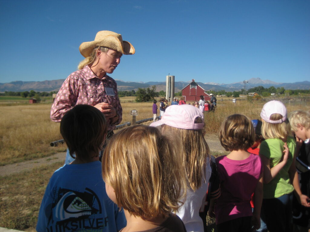 A volunteer speaks to a group of elementary school children during a field trip at the Agricultural Heritage Center. The children face the volunteer, with barns, silos, and mountains visible in the background under a clear blue sky.