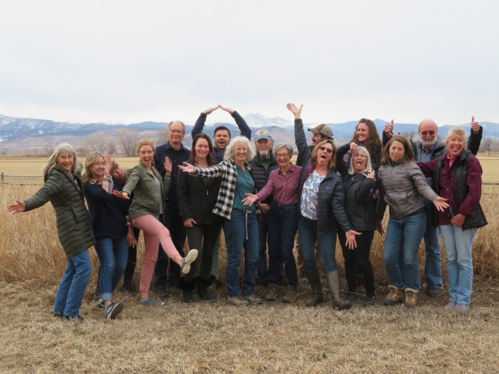 A group of about 16 smiling Volunteer Naturalists pose playfully in an open field with mountains in the background. Some have their arms stretched wide, others make silly gestures, and two people form the shape of a heart with their arms above their heads.