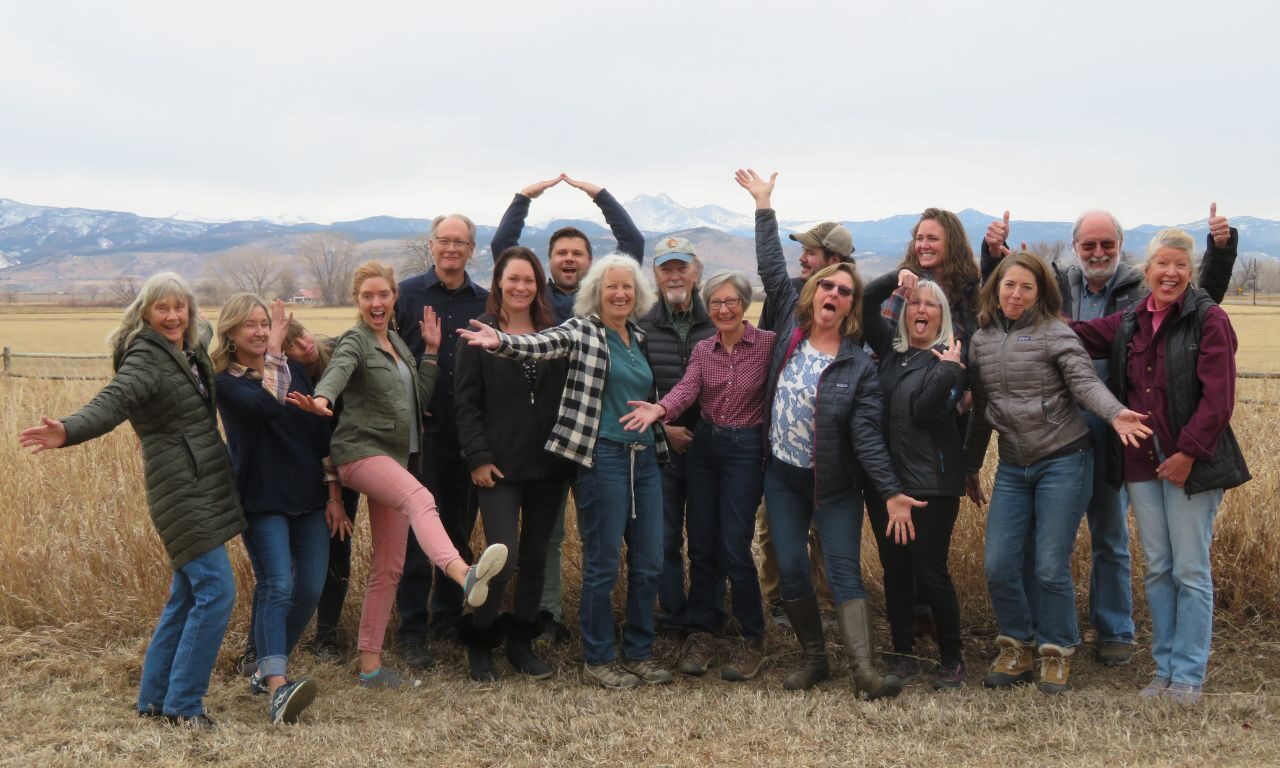 A group of about 16 smiling Volunteer Naturalists pose playfully in an open field with mountains in the background. Some have their arms stretched wide, others make silly gestures, and two people form the shape of a heart with their arms above their heads.